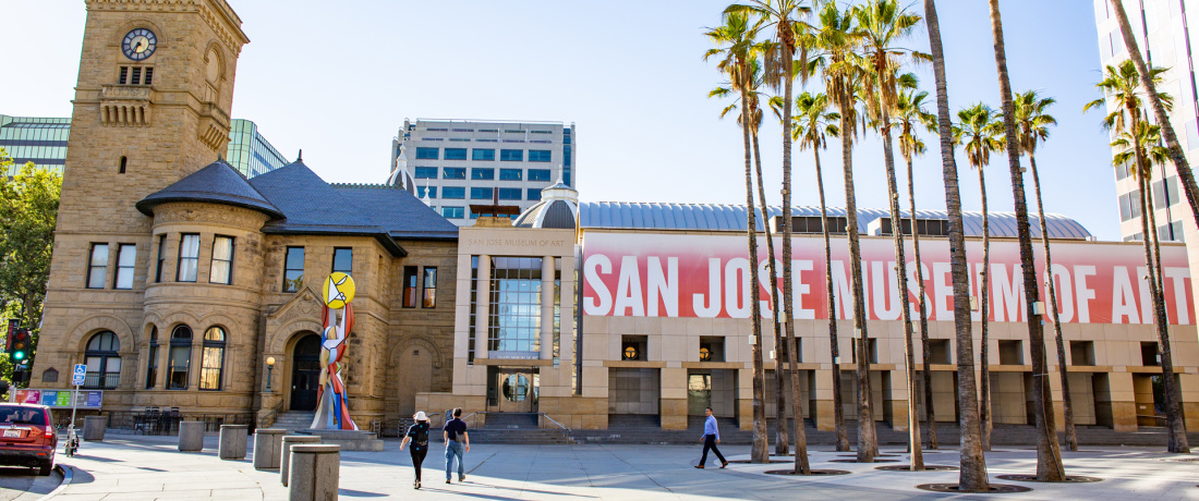 The exterior of a long building that has a large red banner that reads “San Jose Museum of Art.” A circle of palm trees are to the left of the building, with the historic clocktower visible to the far end of the museum. 