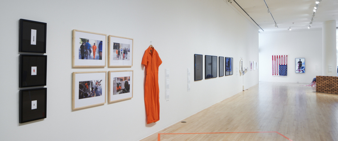 A gallery with an orange prison jumpsuit hangs on the wall, with 3 black framed images to the left of it along with four color framed prints. In the background is an American flag and a partial brick wall with other framed works that are indistinguishable.