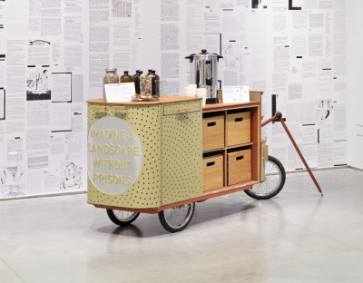 A yellow wooden mobile vendor cart with three wheels sits in the middle of a black and white room. The cart has a door open to reveal drawers, and jars of herbs and a hot water dispenser are placed on top. 