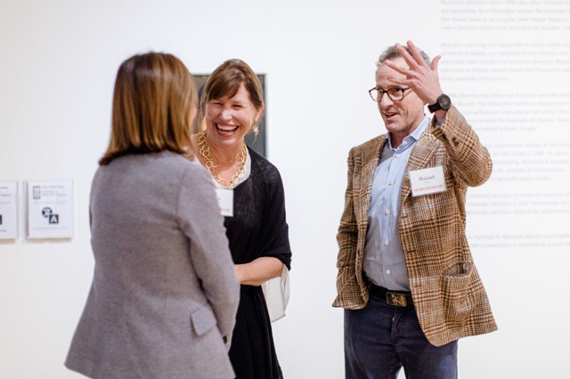 Three people interact in a gallery. Two of them are smiling, one gesturing to the third person.