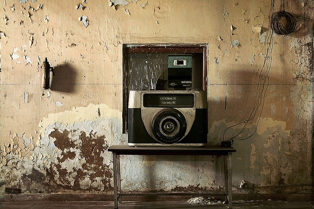 A vintage National 35 Sprinty BC camera sits atop a table in a long-abandoned room. The wall is covered in layers of peeling tan, pale yellow, baby blue, and brown paint—all muted tones.