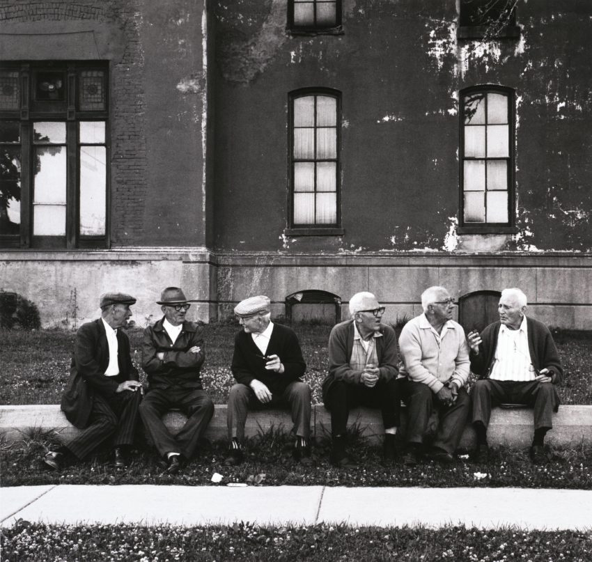 A black and white photograph of 6 elderly men sit on a curb between an aged building and a sidewalk. They are all wearing button ups and slacks, and half of them are wearing glasses and newsie caps. They are engaged in two separate conversations. 