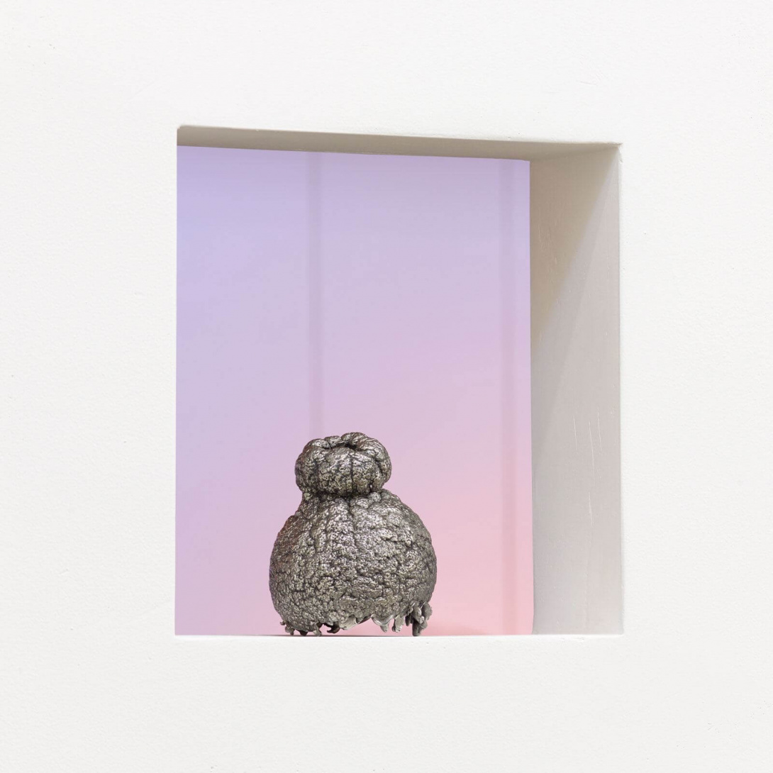 Inside a white framed box is a silver round sculpture. Behind the sculpture is an ombre tone that is pink to lavender.    