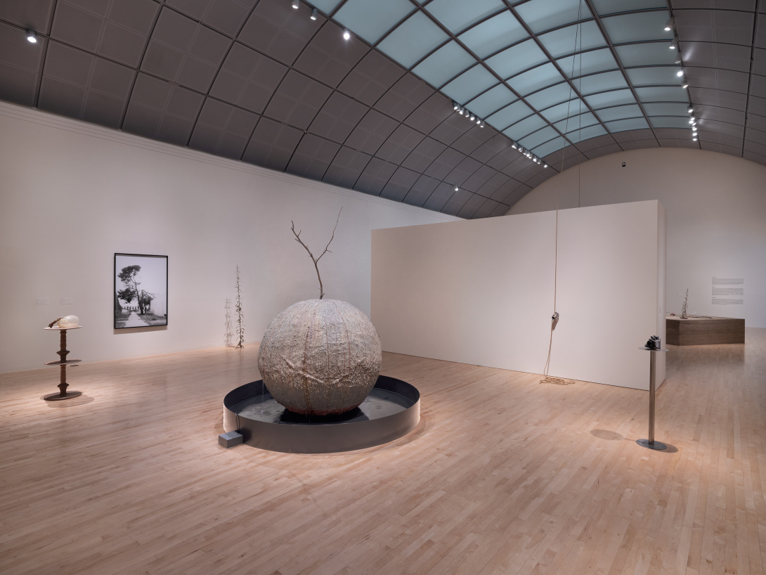 Inside a gallery with an arched ceiling is an oversized round sculpture that sits in a low grey circle. Nearby are thin tall pedestals with small works of art on them. 