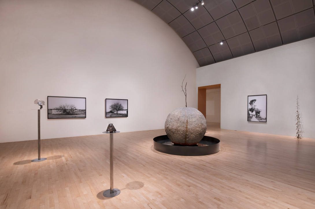 Inside a gallery is a large ball-like sculpture that is nestled inside a low grey circle. In the background are large photographs and throughout the room are thin tall pedestals with tiny sculptures. 