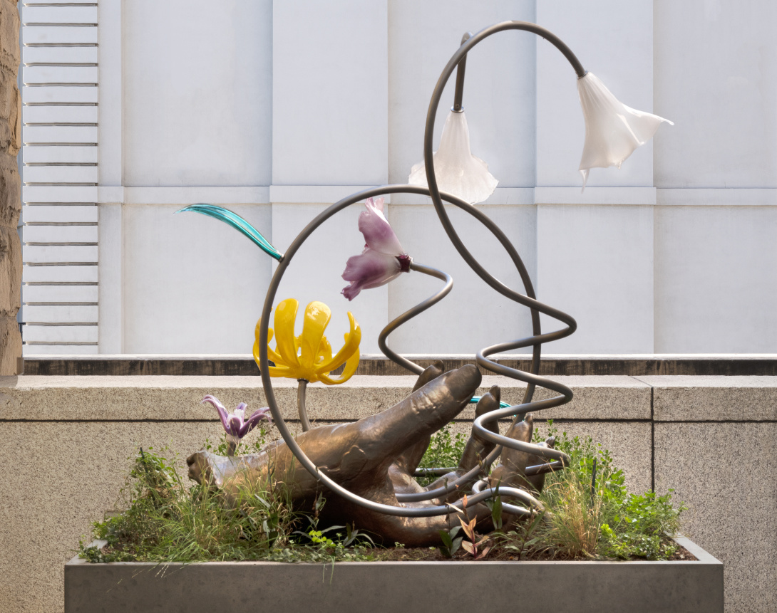 A large outdoor sculpture with flowers hanging and bursting from metal. Living plants grow from the bottom of the sculpture, weaving in and around the sculpture. 