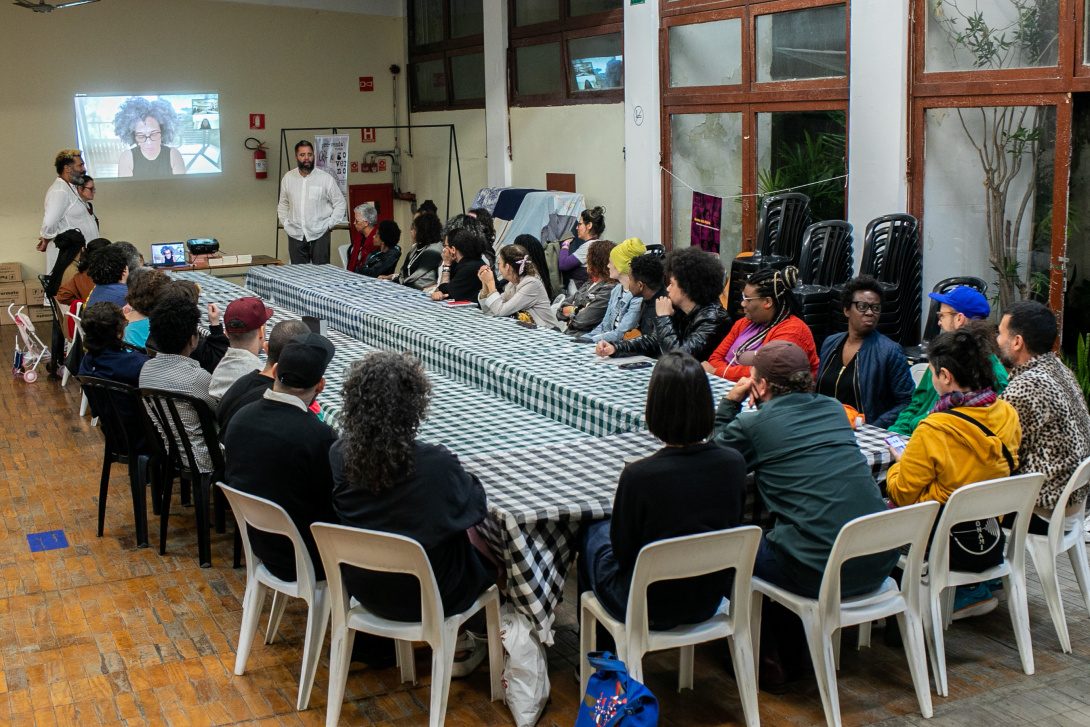 a meeting congregation in a room of twenty people seated together with a person projected on a wall