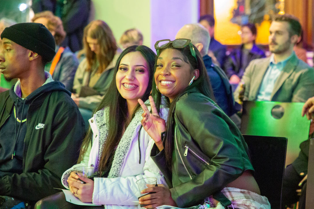Two young women smile and pose for the camera during a seated event at SJMA, with other attendees visible in the background.