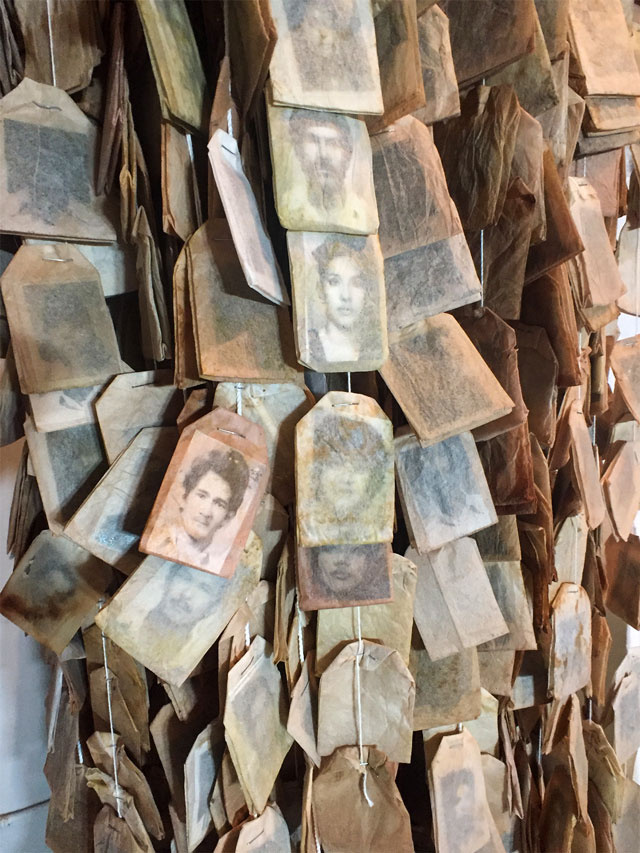 Close up color image of used tea bags containing small black and white photographic portraits. The tea bags are strung together vertically forming a larger mass.