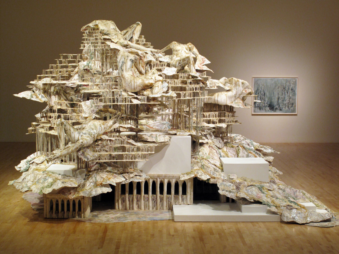 Inside a gallery, a large, multi-layer sculpture appears as an ancient cityscape built into mountains. An unidentifiable white-ish material appears on top of the various layers and structures, acting almost like stretching people, treetops, ceilings, and clouds.