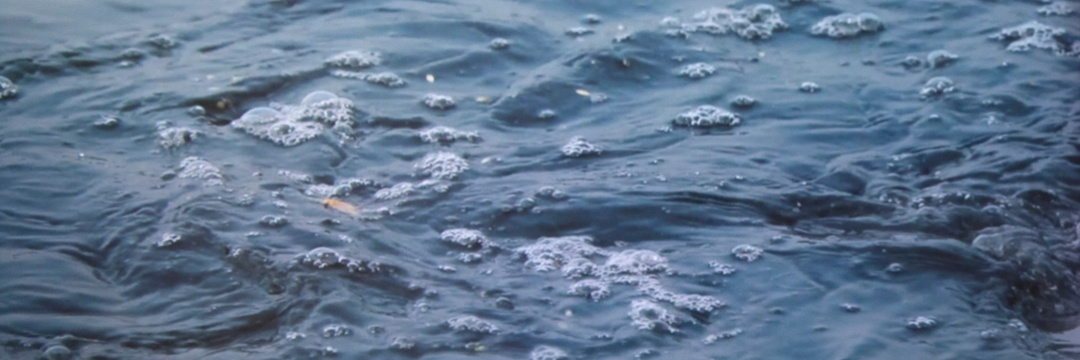 A still image of a water surface shows small splashes of deep blue water. The unknown orange substance is slightly visible both underwater and on top of the surface with bubbles forming around.