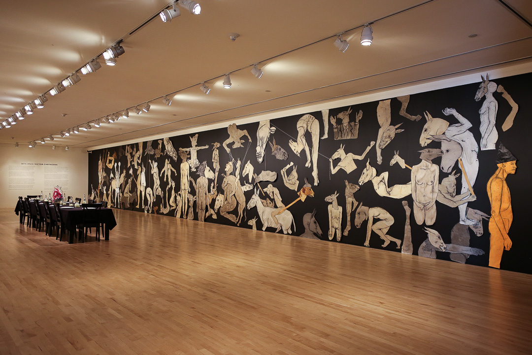 A site-specific installation consisting of a dinner table with place settings and a mural across the length of the gallery wall. The mural depicts life-size human and animal figures in muted colors drawn on paper and set against a black background.