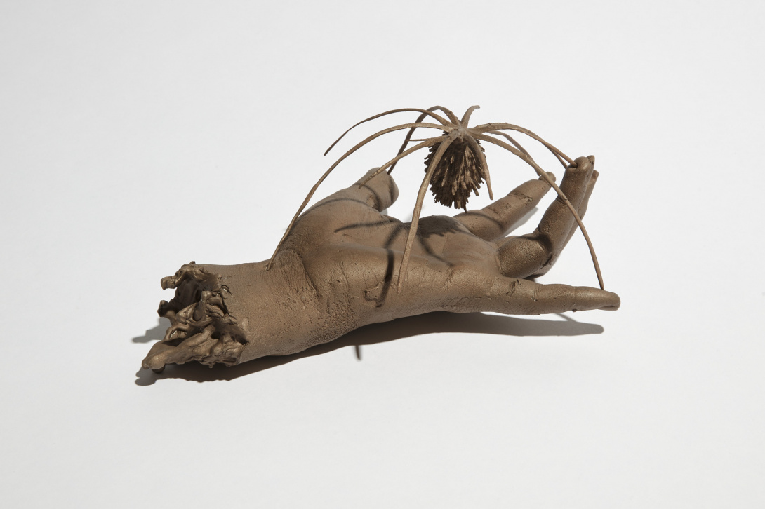 A bronze sculpture of a hand, with open fingers, slightly touching and holding a large plant like object that also resembles a spider. The tendons are visible from the wrist. 