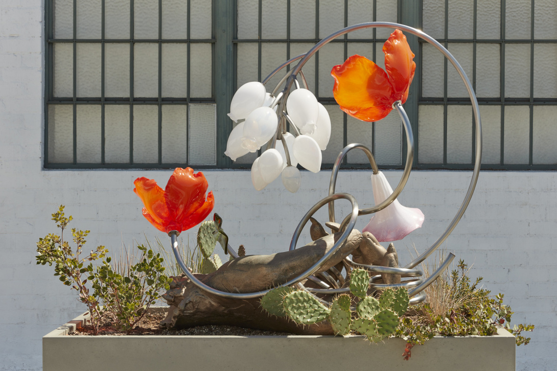 A large outdoor sculpture with flowers hanging and bursting from metal. Living plants grow from the bottom of the sculpture, weaving in and around the sculpture.