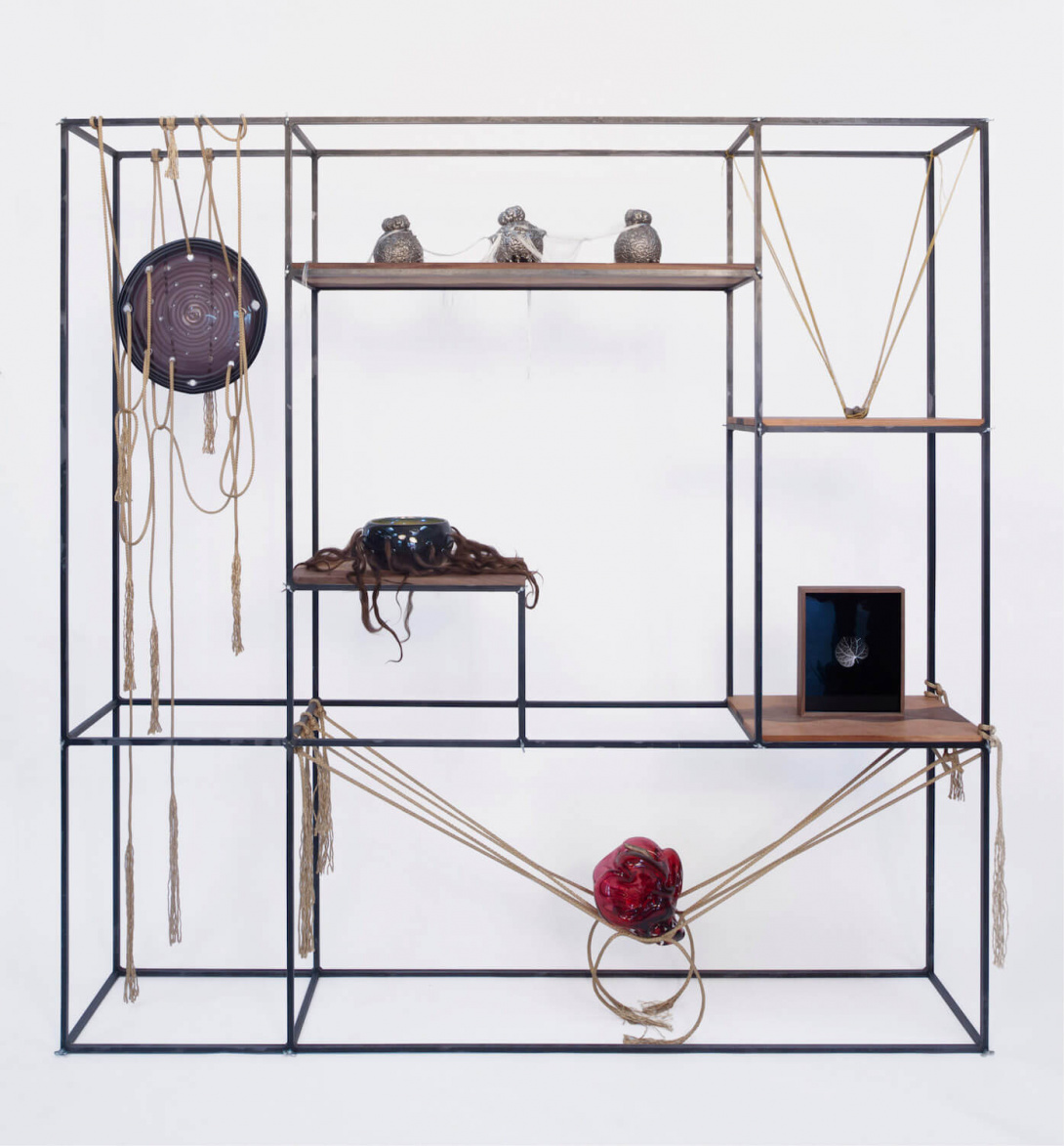 An installation of art objects sit on and hang from a large metal square object. Some objects are round and hard, while others look round or very soft, made of rope.  