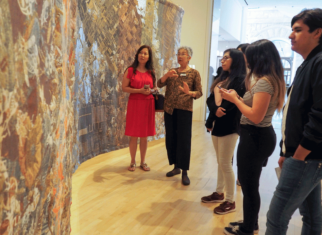 A docent stands in a museum gallery, speaking to a group of high school or college students. The students and professor are looking at a large woven work made of paper or cloth in shades of brown and gold that stands nearly the height of the gallery. 