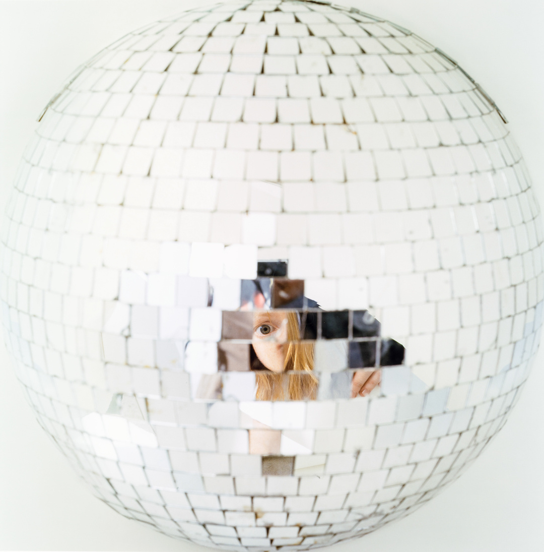 An extreme closeup of a disco ball. Reflected partially in some of the disco ball mirrors is a young red-headed woman taking a selfie with her phone. Her likeness is refracted and visible in a small curvature of the disco ball.