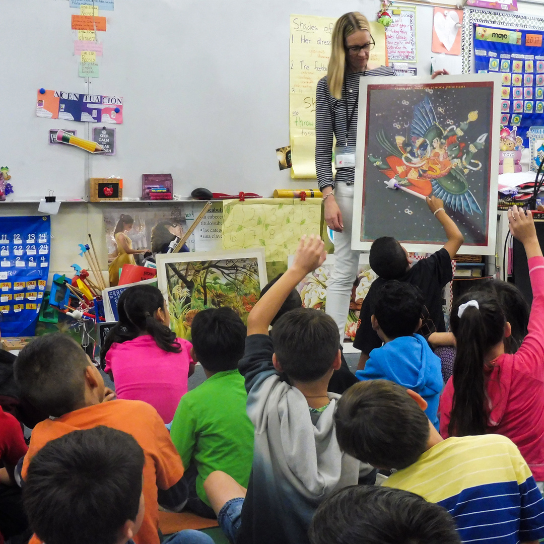 A teacher stands in front of a classroom of young students who are looking at a photo that the teacher is holding. Multiple students sit with their hands raised.