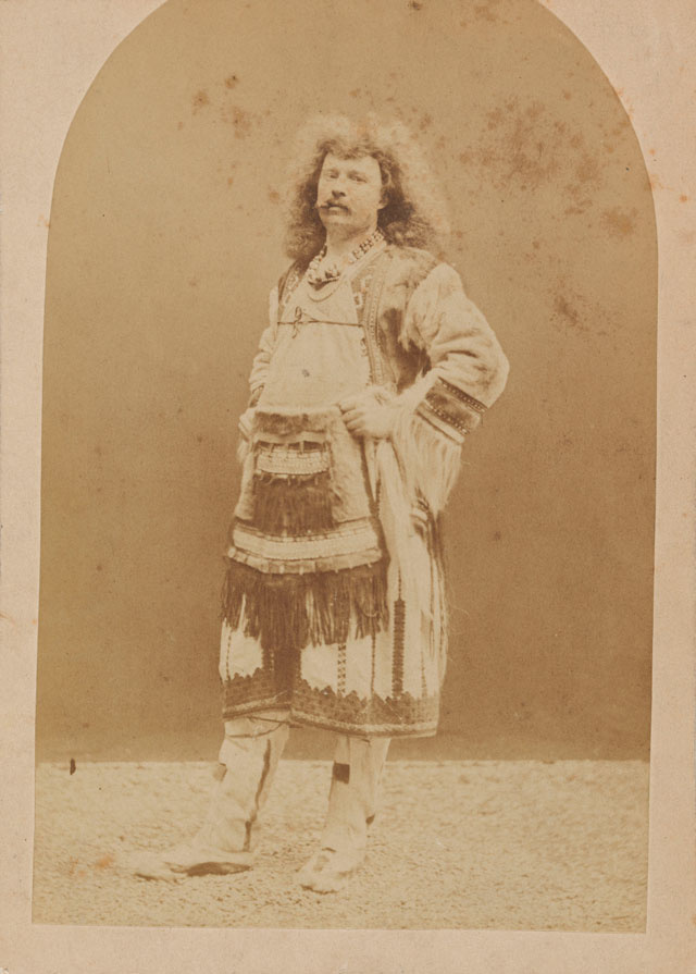 A white man with long frizzy hair, with a cigar in his mouth looks directly at the viewer. He wears Native American clothing consisting of boot moccasins, a fringed leather top and skirt. His arms rest jauntily on his waist, as though daring someone something.