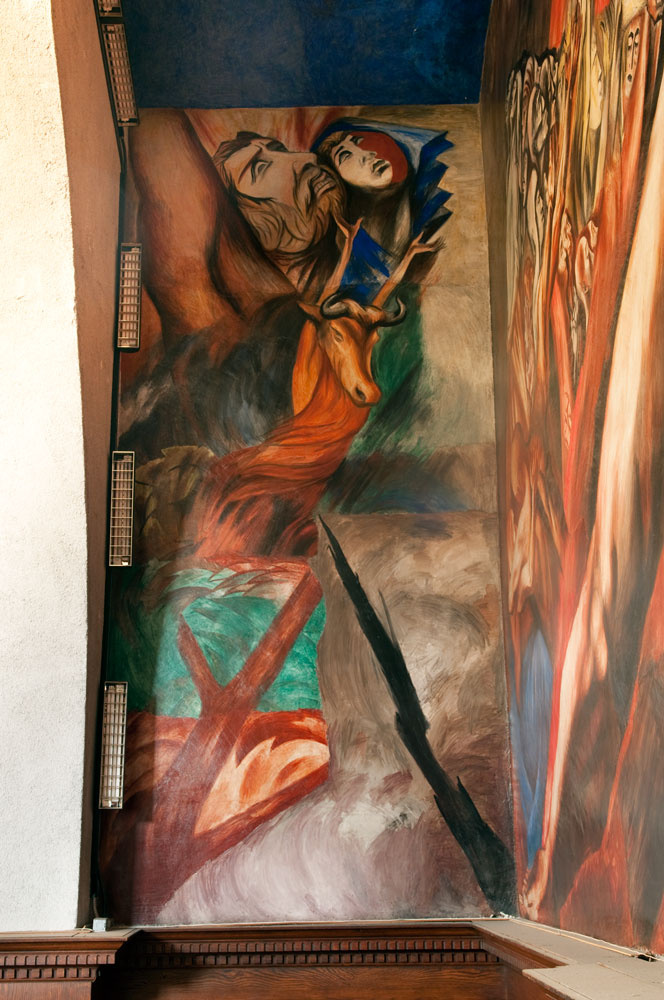 A photograph of a mural installation. One side is an abstraction of two god-like figures in the sky. A creature with arms is pushing up towards them with their hands. To the right is another mural but due to angle, the subject is indecipherable.