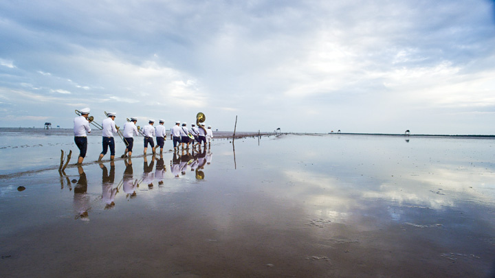 A cloudy sky with muted colors is reflected across a distant horizon line onto an expansive and still body of shallow water. To the left, 10 uniformed figures playing instruments march away in ankle-deep water. They are doubled by their reflections in the water. 