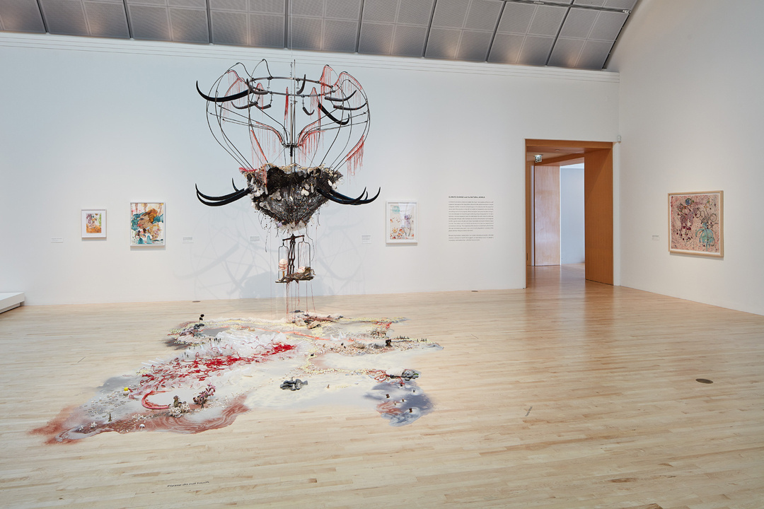 A hanging sculpture in a gallery resembles a bird’s nest attached to a skeleton hot air balloon. Long upward-turning black horns and red tassels adorn it. The gallery floor is covered with white sand peppered with a river of red, patches of blue, and mini bushes or trees.