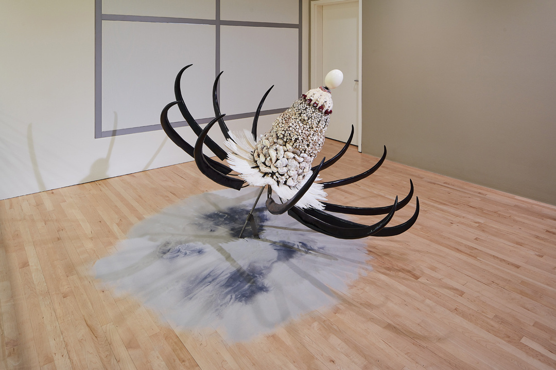 A large sculpture on the floor in the corner of a gallery emerges diagonally out of a shallow pile of white sand on the floor. It resembles something between a satellite disk and an oversized flower and is made of upward curving black horns, shells, and feathers.