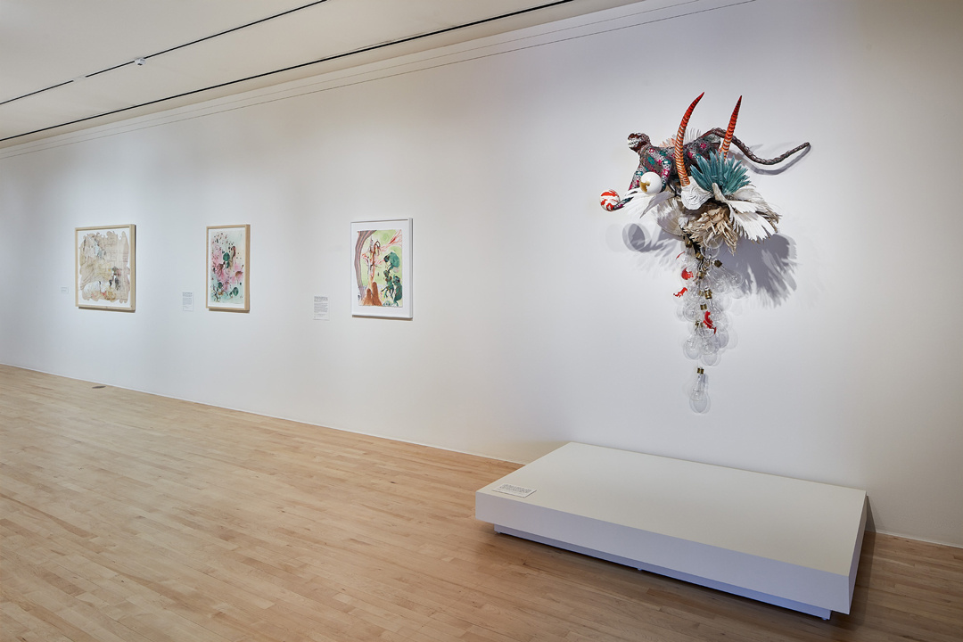 A gallery installation view shows three small framed paintings and a colorful relief sculpture mounted next to them. The sculpture is made of feathers, curving black horns, and other materials including a fantastical reptilian creature holding 2 orbs in its front claws.