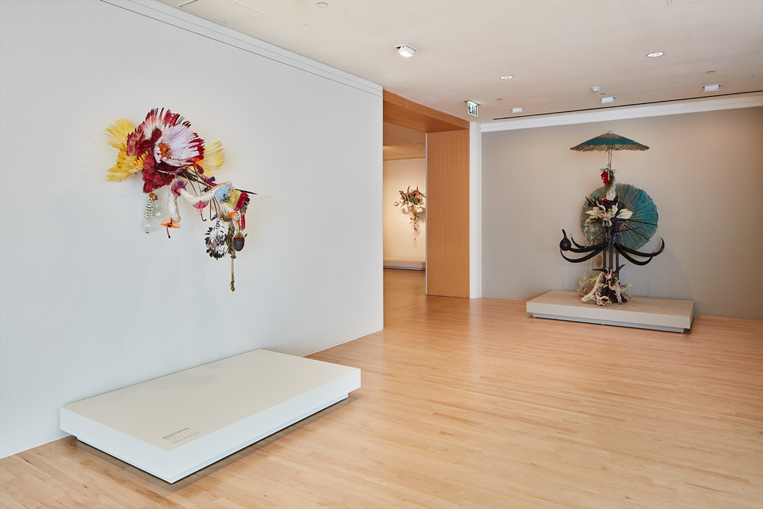 In a gallery corner are 2 multimedia sculptures with another work in the distance. The left is an avian creature with red and yellow feathers, a pointy stick, and other found objects. The right resembles a human holding an umbrella above, made of unknown objects.