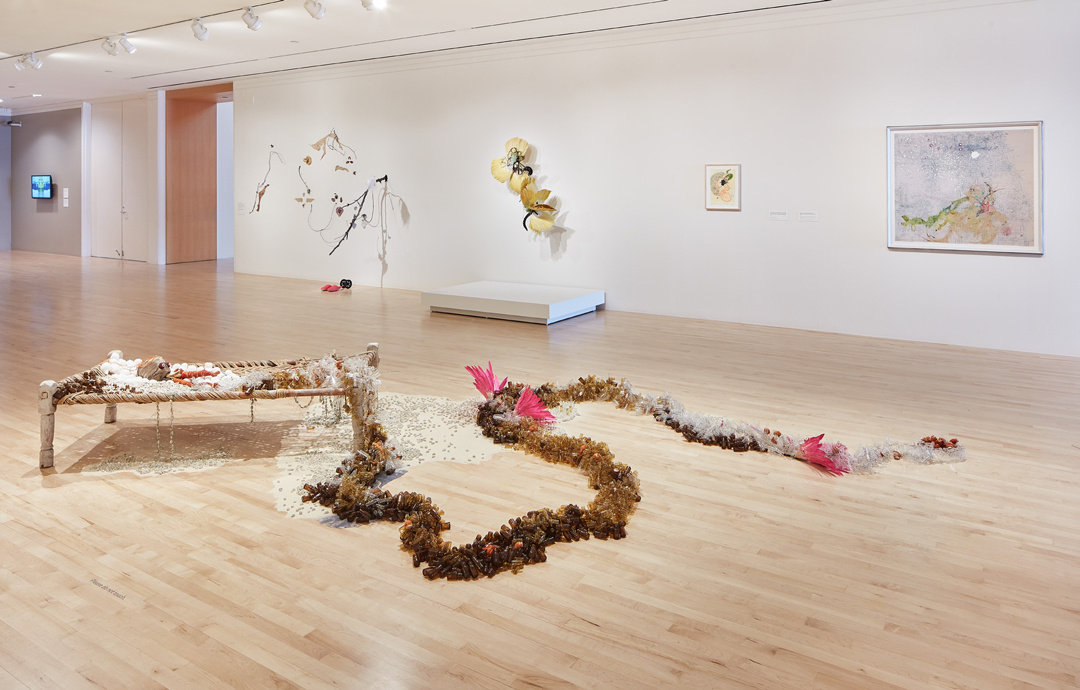 A gallery with a low table in the middle with sand surrounding the table and shell-like items on it. Seaweed appears to be draped around it with more sand. On the wall behind are 2 plant-like sculptures and 2 paintings. In the distance is a screen with indecipherable content.