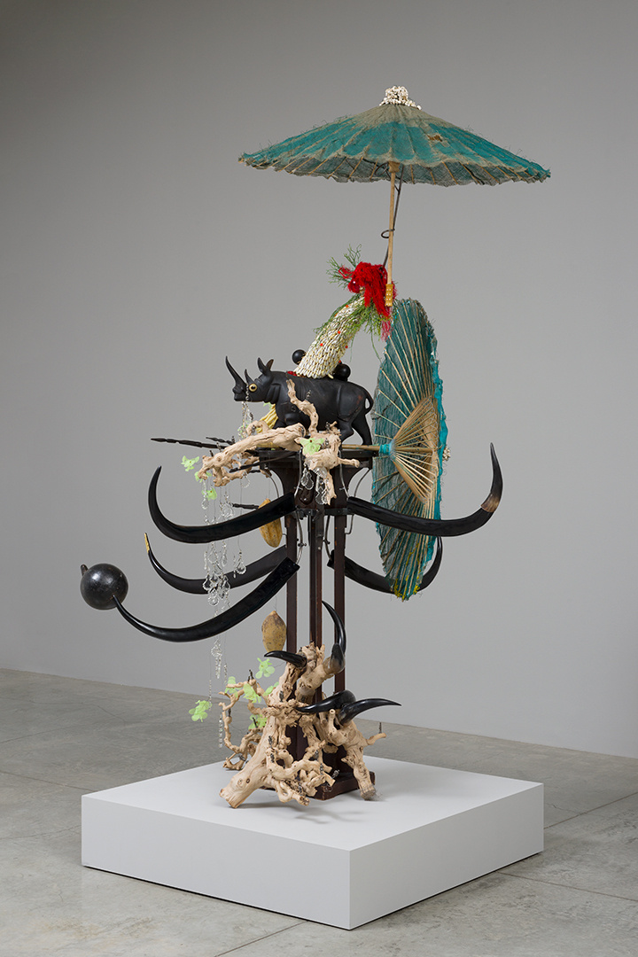 A tall sculptural work sits on a white pedestal, black long horns and tusks protrude from the middle. Drift wood comprises the base, two tattered parasols stick out from the very top and the back, a black rhino figure is nestled in the middle.