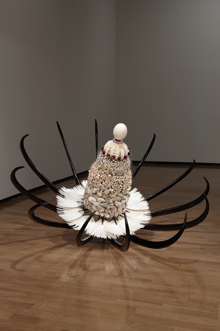 A large flower-like object rests slightly tilted on the floor. The exterior petals resemble black tusks with contrasting white feathers making up the inner petals. The center is a combination of ocean shells with a large white egg sitting at the very top.