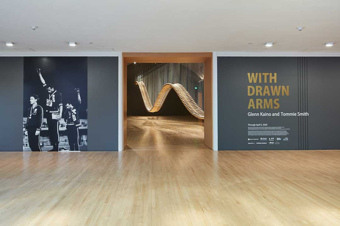 Next to a gallery entrance, a wall reads "With Drawn Arms by Glenn Kaino and Tommie Smith." On the left 3 athletes receive awards while 2 of them hold their arms and fists high. In the gallery is a wave-like object which looks to be made of wooden slats. 