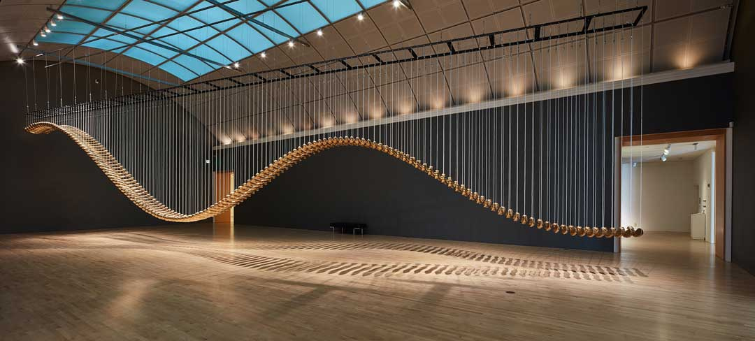 A large gallery filled with a huge structure hanging in mid-air by string-like chords. The sculpture has a lot of movement and looks a bit like a bending spine or a snake moving. It appears to be made of wooden slats.