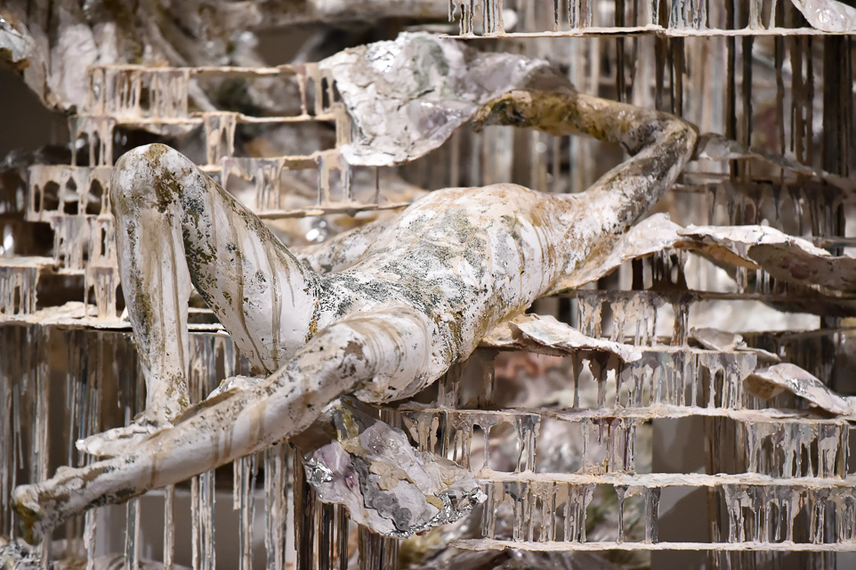 A closeup detail of a large sculpture. Focused on a single figure, laying on its back, made of indeterminate white-ish rusty materials. The figure is surrounded by delicate and strong drippings of this material that also have various brown shades mixed in.