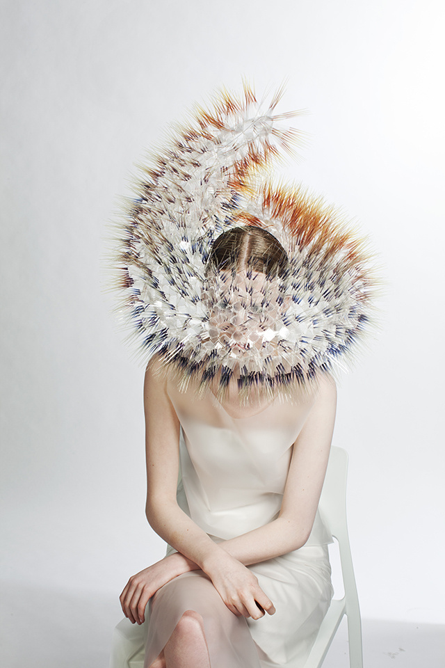 A woman wears a white slip dress is seated in a chair. Her face is obscured by a delicately twisting headdress. The garment comes down over her face and is made up of translucent material. Soft spikes protrude from every inch, akin to a caterpillar, creating the illusion of fur.  