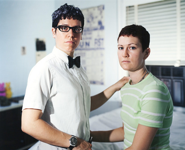 In a blurred bedroom are 2 people with solemn expressions and are slightly holding onto each other. The person on the left wears a white-collard shirt with a bowtie, glasses, a watch, and has slightly sliver hair. The other person wears a striped shirt. 