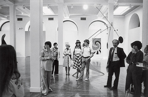 A black and white photograph of a group of people standing in a gallery. They have looks of observation and are talking about something out of the view of the camera.