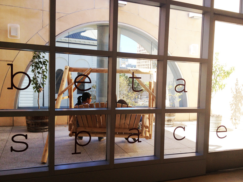 An interior view from behind of two people sitting in a wooden swing in an outdoor, enclosed patio on a sunny day. The photograph was taken through a grid of windows with three rows of windows. Each square window pane has a vinyl letter adhered spelling the words "beta space."