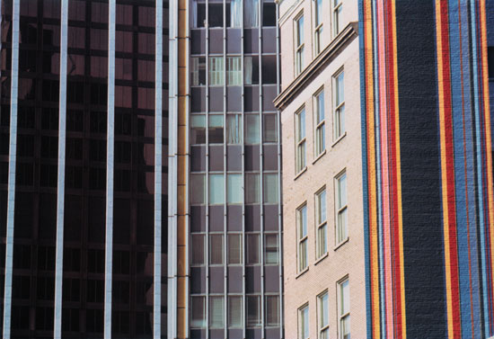 An image or painting of various buildings close together. The first has dark windows reflecting grids of another building, then a grey building with windows. Next to that is a cream building with windows on one wall and a multicolored outside wall facing the viewer.