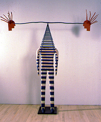 A human shaped sculpture with black and white stripes painted horizontally that go over the body and form a railroad track, becoming smaller the higher up it goes. Balanced at the end is a long black wire that connects to red heads.