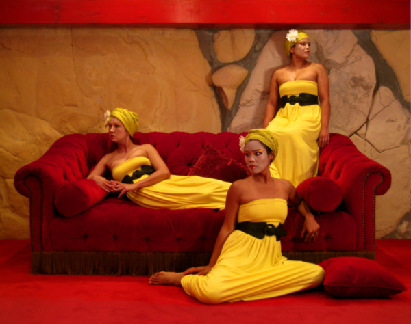 A photograph of three powerful women sitting on and in front of a red velvet couch. They each look in different directions. All are wearing a strapless yellow floor-length dress with a black belt and yellow head wrap.