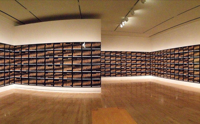 A photograph of a gallery space with 5 walls that each display 6 rows of hanging framed pictures that span from the floor to the ceiling.