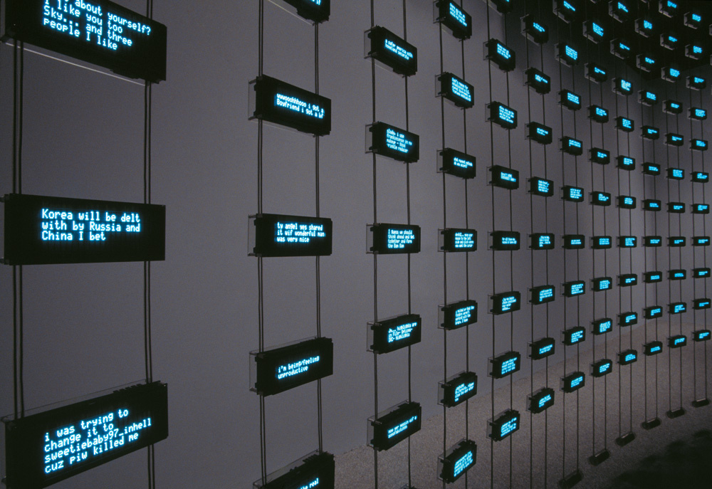 A grid of small rectangular machines suspended in the air. The room is dark, the only light comes from the LCD text displayed on the screens of these small machines. The words appear to be fragments of conversations.