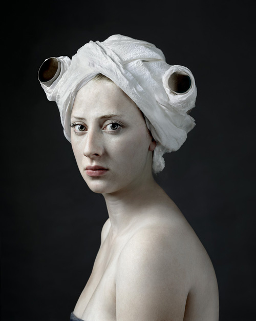 A photograph of a bare shouldered female with head wrapped in toilet tissue and paper rolls used as rollers. The woman's face is solemn with pink lips and long eyelashes. She is set against a dark background.