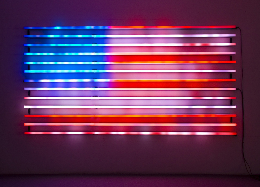 A digital light sculpture mounted on a wall with two cables hanging down the right edge of the photograph. Thirteen horizontal light stripes (or bands) make up an abstracted image of the American flag. The lights glow and reverberate off each other in the darkness.