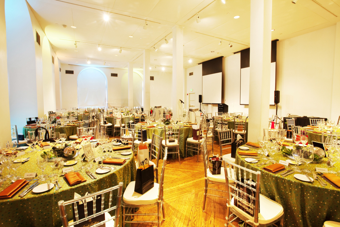 Nearly a dozen round tables with bright green tablecloths stand in a banquet hall. The tables are set for 10 people, with silver chairs, shiny burnt-orange napkins, and gift bags with orange tissue paper inside. 