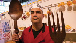 A color photograph of a middle aged male wearing a chef hat standing in front of a display of normal sized kitchen utensils holding an oversized wooden fork and spoon.