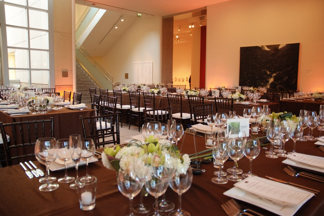 Four long rectangular tables with black table cloths are in a lobby. On them are a variety of wine and champagne glasses with bouquets of white flowers.