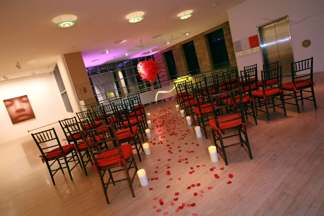 Overlooking a balcony on the second floor, rose petals and lit white candles are on the ground leading down an aisle between rows of black chairs with red cushions. In the background hanging from the ceiling is a red glass chandelier.